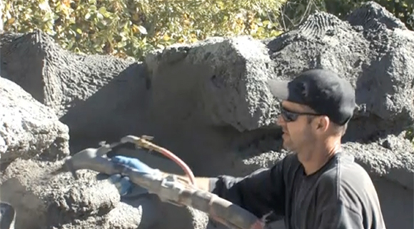 Richard using a pneumatic carving tool to detail and texture a concrete rock surface outdoors