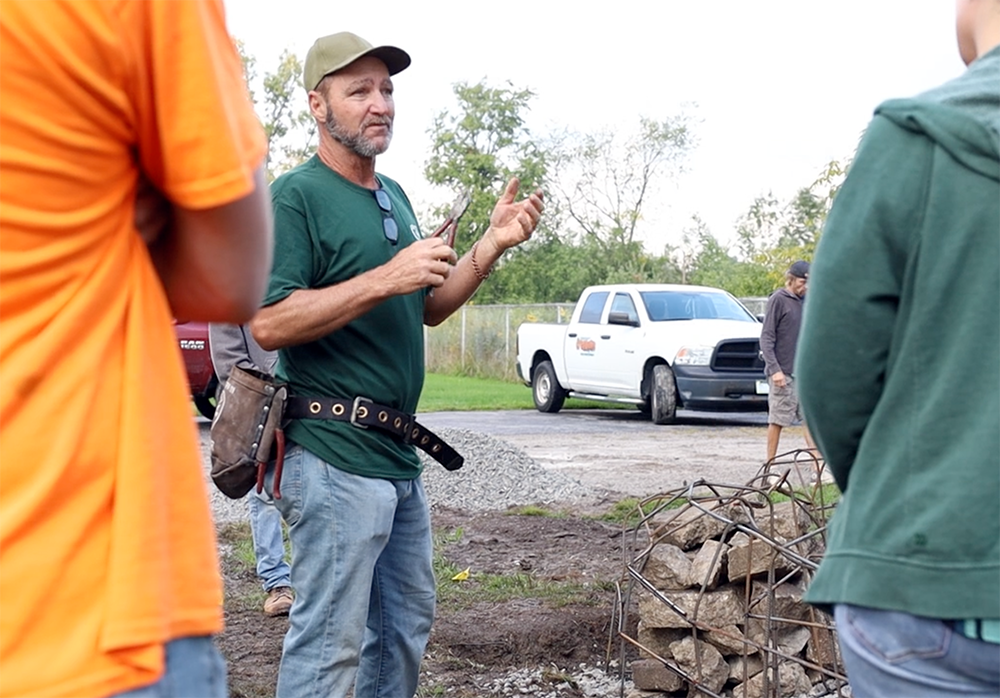 Richard consulting at a construction worksite wearing a tool belt and holding a tool in his hand. Other people stand in the foreground listening to his direction