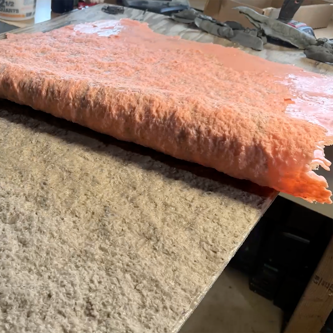 A sheet of orange-coated fiberglass or foam material being peeled back from a textured concrete or stone mold surface on a workbench