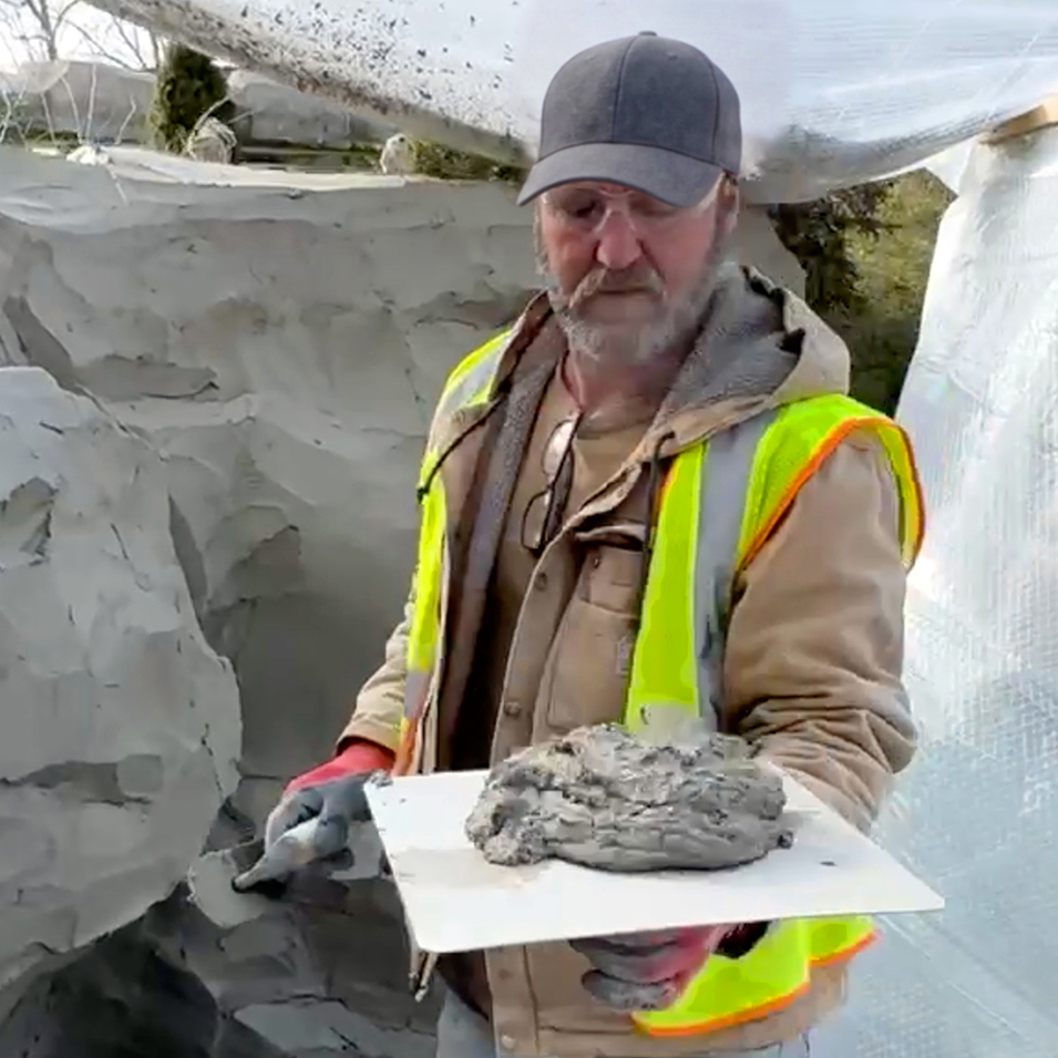 Richard L. Winget in a safety vest and cap holding a plasterer's hawk with a mound of wet concrete mix, standing beside a large sculpted rock form under a plastic enclosure
